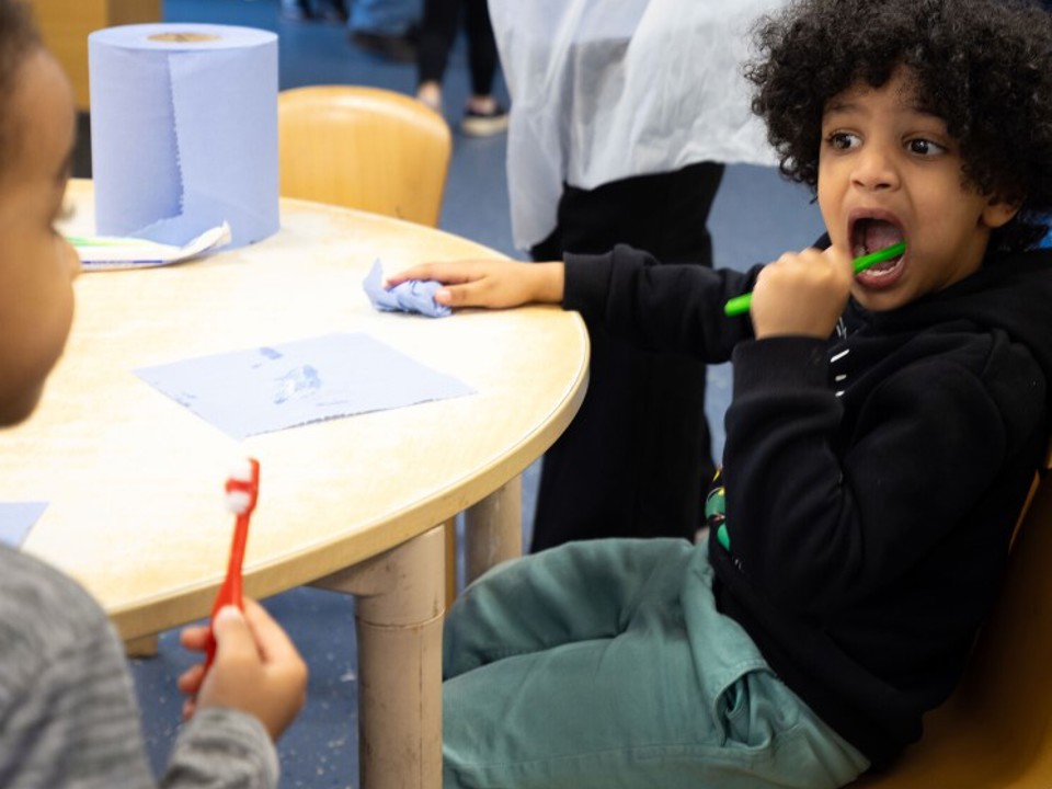 Two young children sit at a round table with blue paper towels on it. One child uses a green toothbrush, appearing focused. The scene is light-hearted.