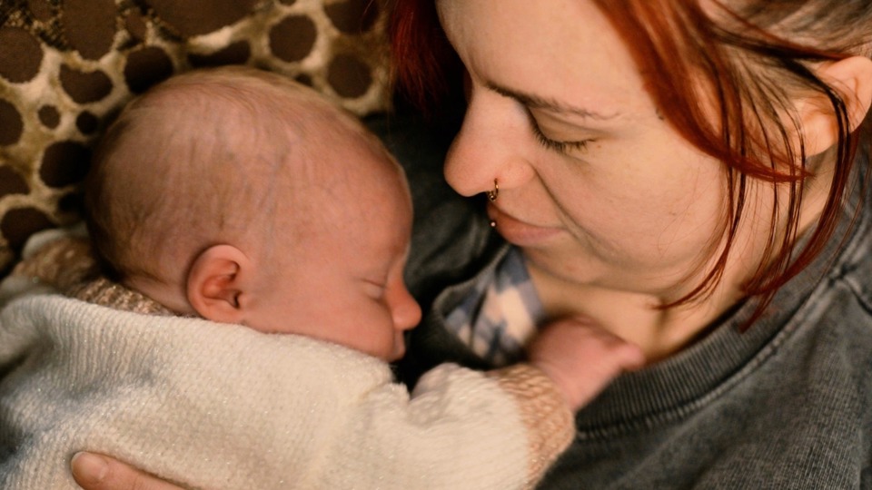A tender moment captured as a woman with light brown hair and a nose piercing cuddles a sleeping baby on her shoulder. Warm lighting, cosy setting.
