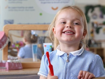 A smiling child in a blue checkered dress holds a red toothbrush, standing by a mirror with dental models and toothpaste, exuding joy and playfulness.