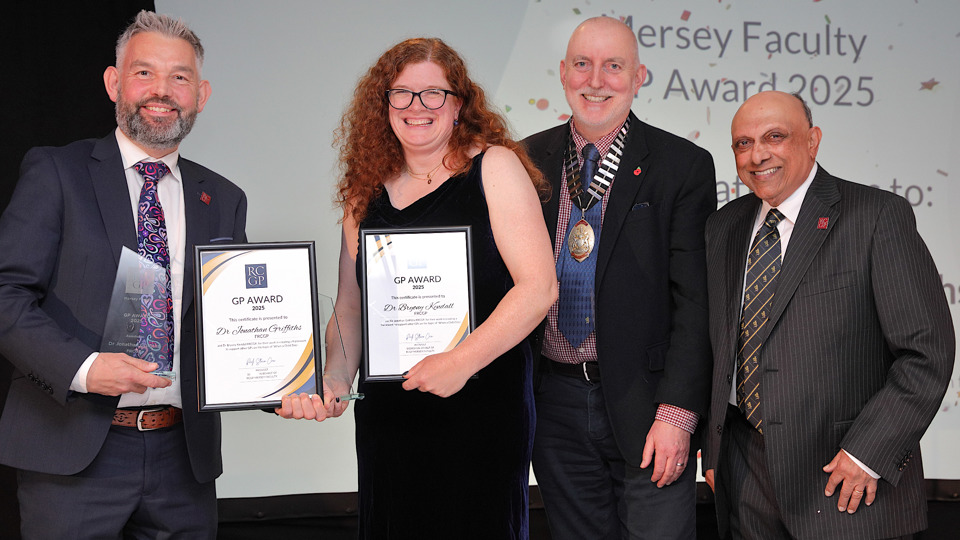 Four people smile at an award ceremony, with two holding certificates. The background displays "Mersey Faculty GP Award 2025." They appear celebratory and formal.