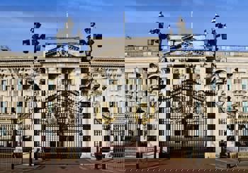 Elegant view of Buckingham Palace's grand facade and ornate black gates, adorned with golden crests, under a clear blue sky.