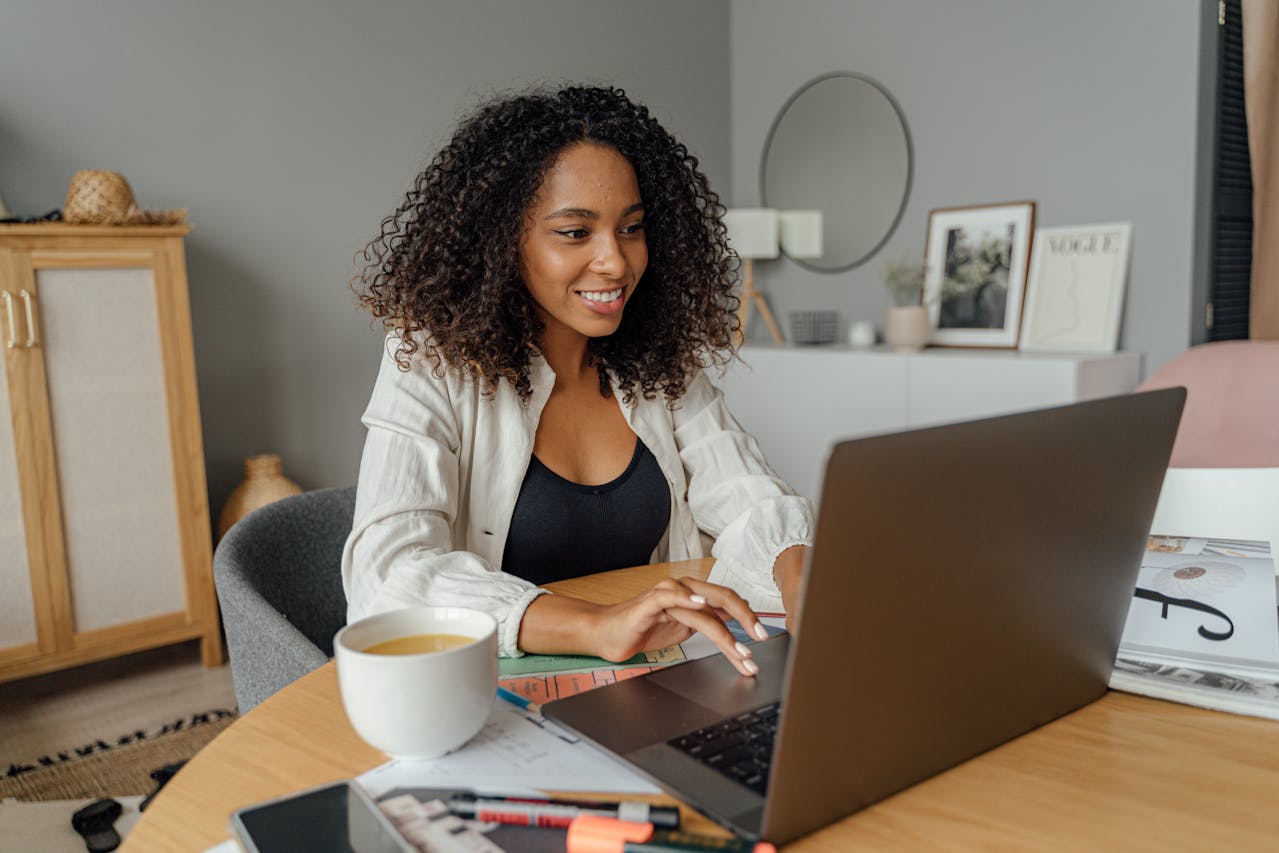 Smiling woman with curly hair works on a laptop at home. She's seated at a wooden table with a coffee mug, papers, and stationery, creating a cosy, focused ambiance.