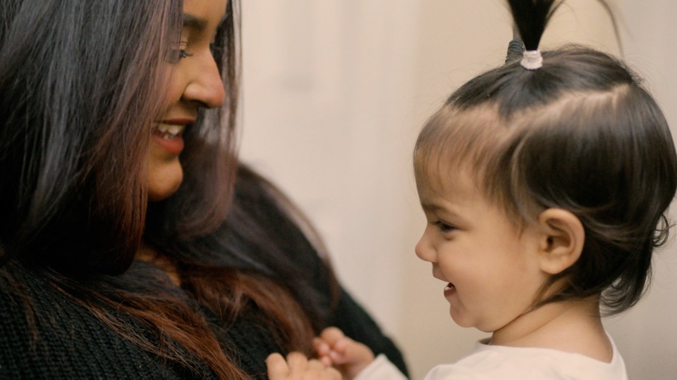 A smiling woman with long hair looks at a cheerful toddler with a small ponytail. They are facing each other, conveying warmth and joy in a close bond.