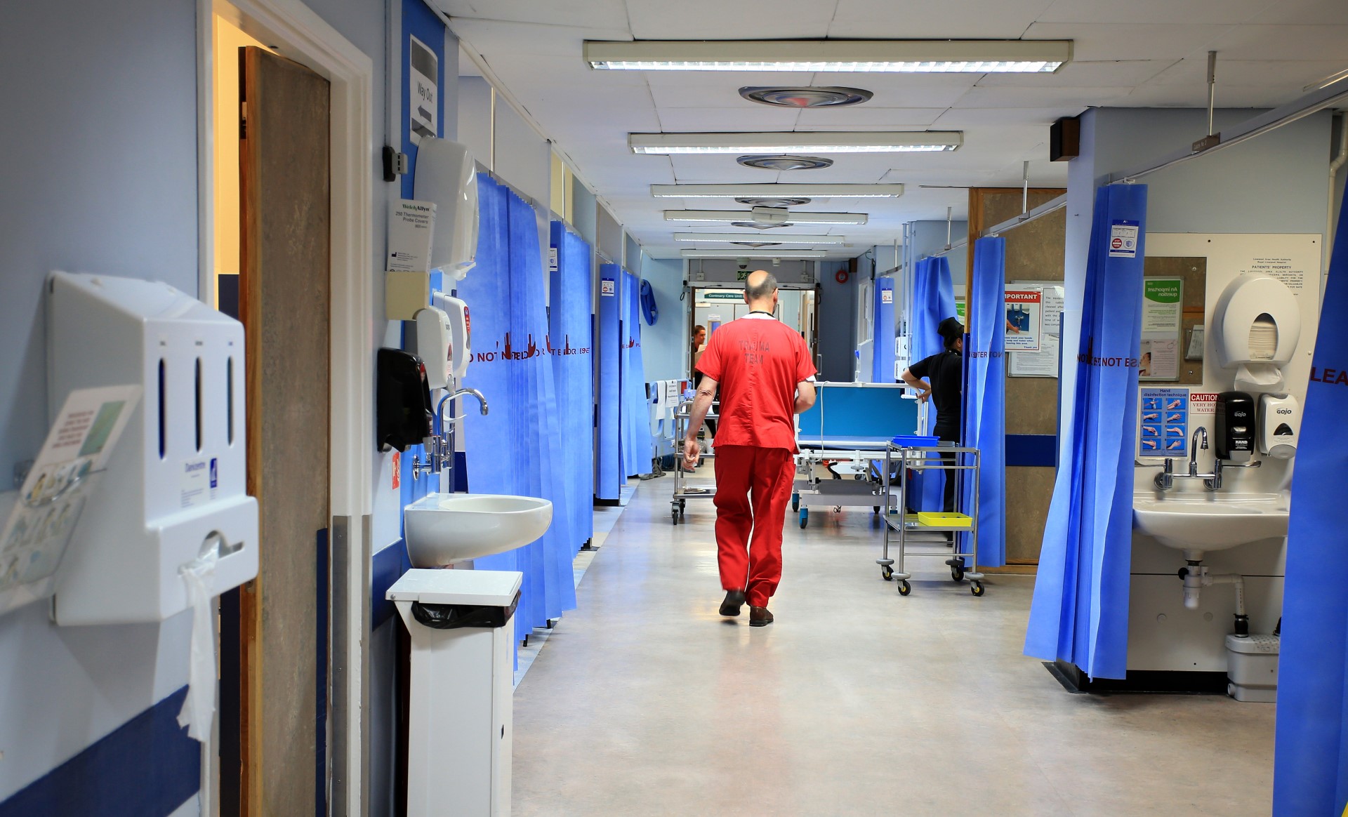 Hospital corridor with blue curtains and medical equipment. A healthcare worker in red scrubs walks down the hallway.